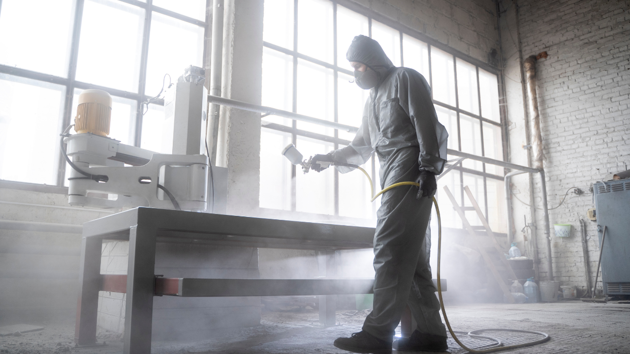 Trabalhador aplicando pintura em spray em uma peça metálica em um galpão industrial, usando macacão e máscara.